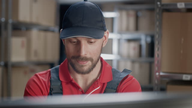 Portrait-of-Uniformed-Worker-Using-Personal-Computer-while-Sitting-at-His-Desk-in-the-Warehouse.-In-the-Background,-Room-with-Shelves-Full-of-Cardboard-Box-Packages-Ready-For-Shipping.