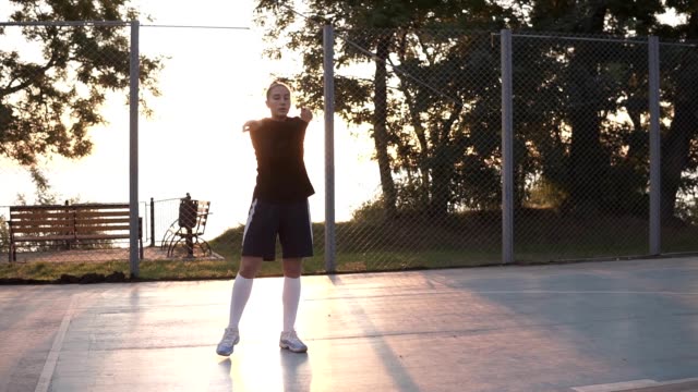 Caucasian-sporty-young-woman-in-hoodie-and-shorts-warming-up-in-morning-on-basketball-court.-Sun-shines-on-the-background