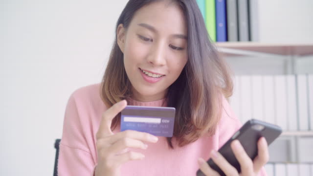 Hermosa-mujer-asiática-utilizando-smartphone-comprar-online-compras-con-tarjeta-de-crédito-mientras-usar-suéter-sentada-en-el-escritorio-en-la-sala-de-estar-en-casa.-Mujer-de-estilo-de-vida-al-concepto-de-hogar.