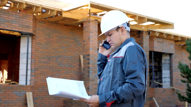 Hombre-trabajo-con-el-proyecto-de-papel-tener-llamada-de-teléfono-permanente-en-el-exterior-del-sitio-de-construcción-de-casa