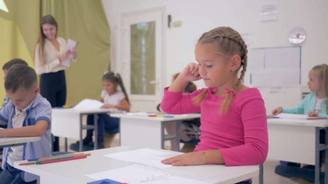 Porträt-von-Schulmädchen-auf-Zeichnung-Lektion-in-helle-Klassenzimmer-sitzen-hinter-Schreibtisch-auf-Hintergrund-von-Klassenkameraden-und-junge-Lehrer