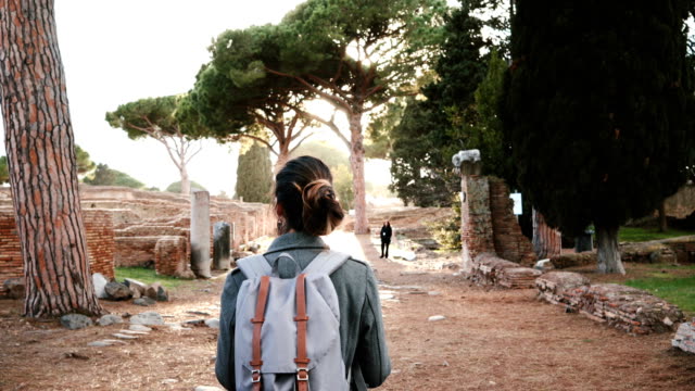 Back-view-of-beautiful-tourist-girl-with-backpack-and-map-exploring-ancient-sunny-amphitheater-ruins-in-Ostia,-Italy.