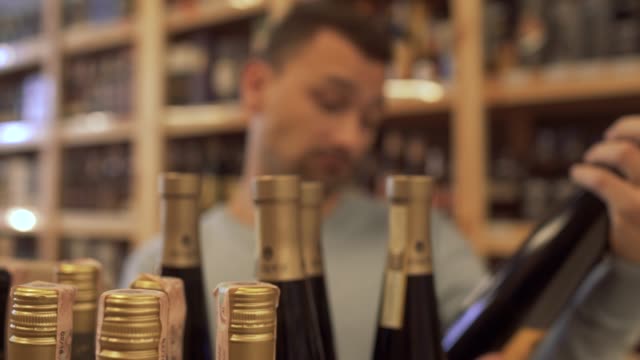 Customer-reading-the-composition-of-the-wine-on-the-bottle-label,-nods-approvingly-and-going-away-taking-the-wine-bottle-with-him.-Guy-who-choosing-the-wine-in-the-store-is-out-of-focus.