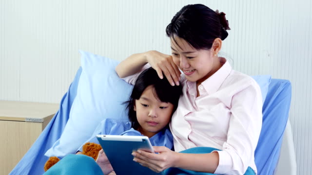Little-girl-and-her-mom-using-tablet-together-at-hospital.-People-with-Technology,-Family,-Healthcare-and-Medical-Concept.