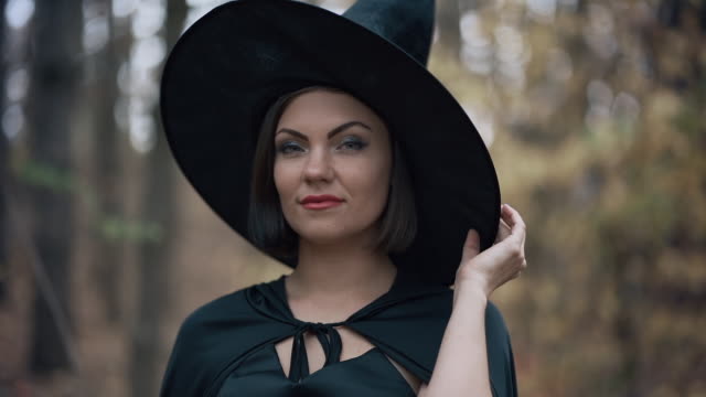Portrait-of-young-pretty-witch-in-cap-on-autumn-forest-background.-Halloween-concept,-cosplay-dressing-up.-Slow-motion