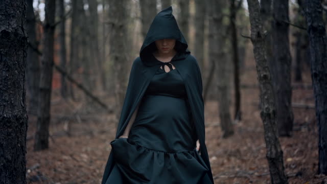 Black-witch-in-hood-walks-in-autumn-scary-forest.Womanl-in-long-dress.-Halloween,-horror,-concept,-cosplay-dressing-up