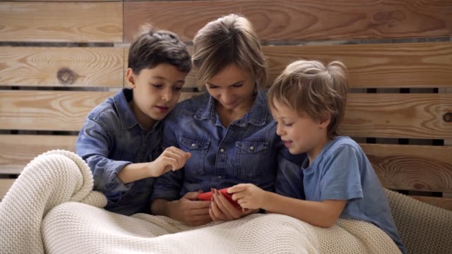 Joven-mujer-caucásica-con-dos-hijos-se-divierten-usando-el-teléfono-inteligente-relajarse-en-el-sofá,-los-niños-felices-disfrutan-de-pasar-tiempo-en-casa-con-mamá.-Videojuegos