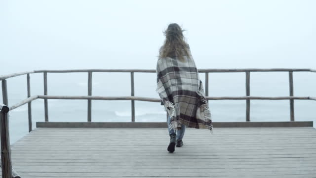 Back-view-of-young-woman-with-plaid-standing-on-the-wooden-pier-and-looking-afar.-Female-spends-time-alone-in-foggy-day