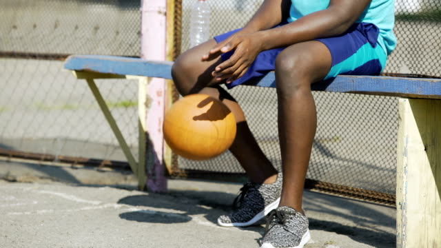 Close-up-of-African-American-male-sitting-on-the-bench-and-playing-ball,-sadness