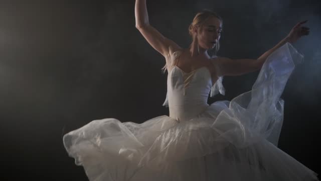 Mid-Shot-of-a-Beautiful-Young-Ballerina-Spinning-Gracefully-in-the-Spotlight.-She-dressed-in-white-airy-dress-modern-tutu.-Sensual-woman-on-smoke-black-stage.-Slow-motion