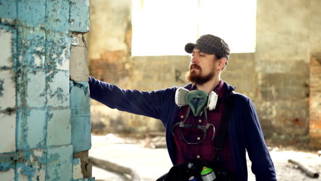 Ernsten-bärtigen-Mann-konzentriert-sich-auf-Graffiti-auf-alte-schmutzige-Spalte-in-verlassenen-Gebäude-mit-Aerosol-Farbe-malen.-Mann-ist-legere-Kleidung-und-Handschuhe-tragen.
