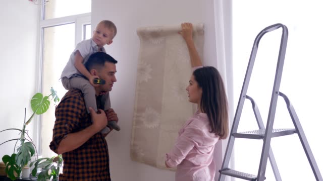 glückliche-Familie-Reparaturen-im-neuen-Heim,-fröhliche-Mama-und-Papa-mit-Sohn-auf-Schultern-wählte-Tapete-in-Wohnung