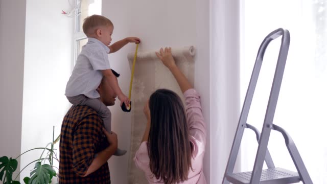 family-renovation-in-room,-young-mother-and-father-with-boy-on-shoulders-glue-new-wallpaper-on-wall