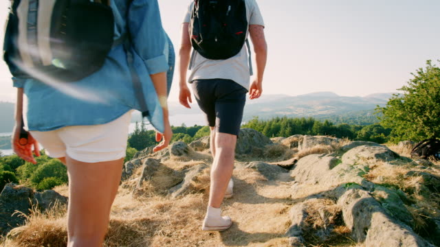 Slow-Motion-Rear-View-Shot-Of-Young-Couple-Standing-At-Top-Of-Hill-Looking-At-Beautiful-Countryside-On-Hike-Through-Lake-District-In-UK