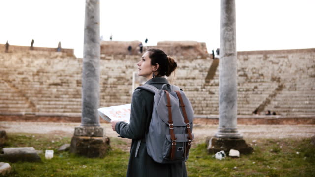 Back-view-of-beautiful-young-female-tourist-with-backpack-and-map-exploring-ancient-amphitheater-ruins-in-Ostia,-Italy.