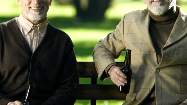 Smiling-aged-men-showing-beer-bottles,-sitting-on-bench,-non-alcoholic-beverage