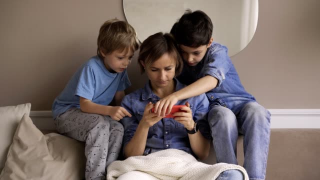 Joven-mujer-caucásica-con-dos-hijos-se-divierten-usando-el-teléfono-inteligente-relajarse-en-el-sofá,-los-niños-felices-disfrutan-de-pasar-tiempo-en-casa-con-mamá.-Videojuegos