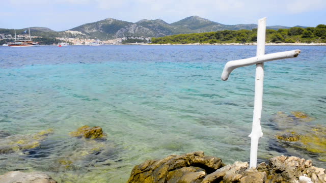 Wooden-christian-white-cross-on-rocky-shore-of-blue-sea.-Seacoast-of-Hvar-island-and-harbour-in-old-town-Hvar.