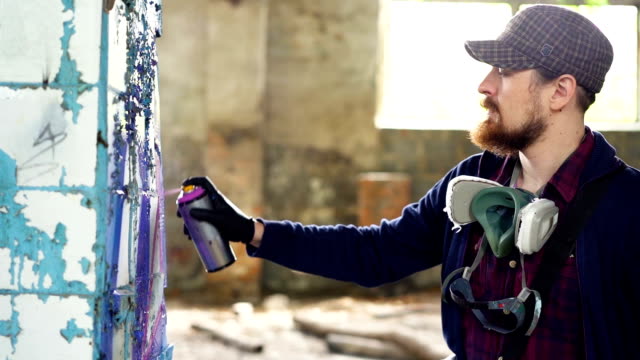 Ernster-Mann-ist-Graffiti-auf-schmutzigen-Säule-in-alten-leeren-Haus-mit-Aerosol-Farbe-malen.-Kerl-trägt-legere-Kleidung-und-trendige-Cap,-er-hat-schützende-Handschuhe-und-Maske.