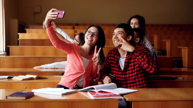 Fröhlichen-Studenten-nehmen-Selfie-im-Hörsaal-sitzen-zusammen-an-Schreibtischen-und-halten-Smartphones.-Moderne-Technologie,-Selbstporträt-und-Bildung-Konzept.