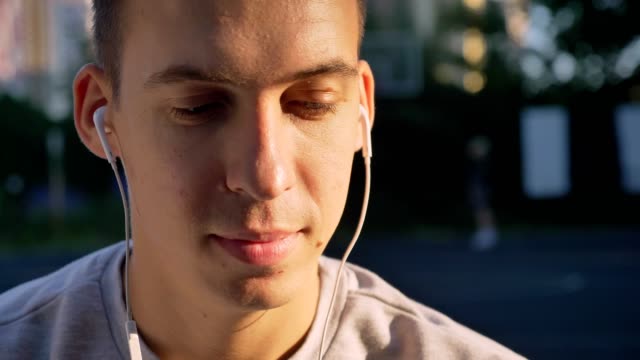 Portrait-of-handsome-young-man-listening-music-through-earphones-and-looking-at-camera,-people-playing-in-background-open-air