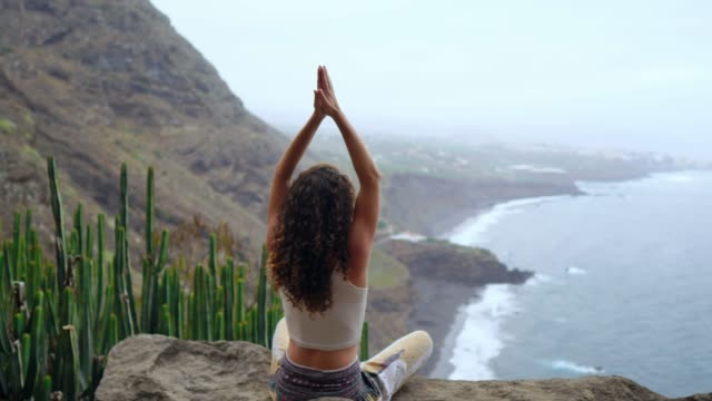 Mujer-joven-haciendo-yoga-en-la-montaña-en-una-isla-con-vista-al-mar,-sentada-en-una-piedra-en-la-cima-de-una-montaña-meditando-en-posición-de-loto