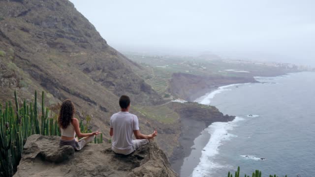 Ein-Mann-und-eine-Frau-sitzen-auf-einem-Berg,-der-Blick-auf-den-Ozean,-sitzt-auf-einem-Stein-in-einem-Lotussitz-meditieren.-Die-Ansicht-von-hinten.-Kanarische-Inseln