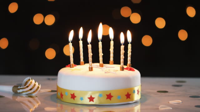 Seven-lit-candles-on-a-white,-decorated-birthday-cake,-a-party-blower-beside-it,-bokeh-lights-in-the-background
