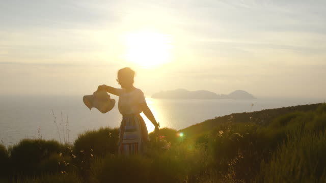 Schöne-junge-Frau-tragen-bunte-Kleid-Mode-mit-Rock-und-Hut-Stand-mit-Blumen-im-Korb-bei-Sonnenuntergang-auf-der-italienischen-Insel-Ponza-Berg-Küste.