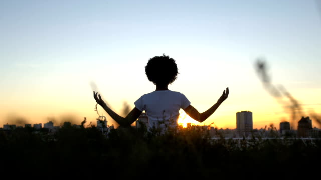 Señora-afroamericana-practicando-yoga,-sentado-en-posición-de-loto,-puesta-de-sol-en-la-ciudad-de