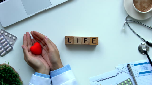 Life-word-made-of-wooden-cubes-on-table,-doctor-hands-showing-toy-heart-top-view