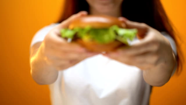 Chica-mostrando-la-hamburguesa-de-carne-de-res-a-cámara,-tentación-por-la-comida-rápida,-poco-saludable-de-comer
