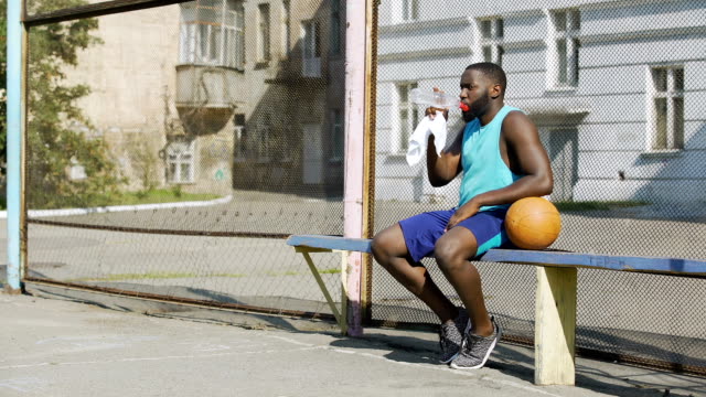 Jugador-de-baloncesto-fuerte-sentado-en-la-banca-y-beber-agua-mineral,-deporte