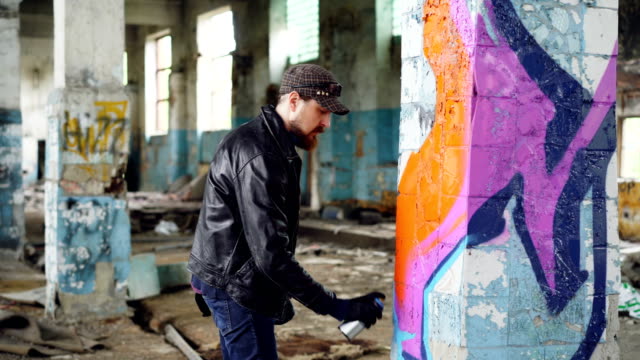 Handsome-bearded-guy-graffiti-artist-is-painting-with-spray-paint-inside-abandoned-building.-Modern-street-art,-youth-subculture-and-creative-people-concept.