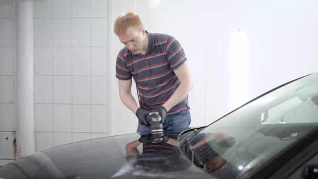 redhead-male-worker-is-processing-surface-of-car-by-polishing-machine-in-a-car-service,-covering-by-protective-layer