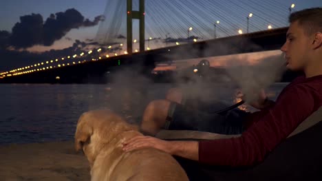 Junger-Mann-mit-einem-Hund-am-Strand-sitzt-und-eine-Wasserpfeife-raucht