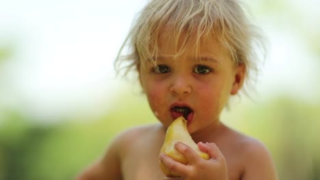 Ehrliches-Porträt-von-blonde-Baby-Kleinkind-Essen-Birnenfrucht-im-Freien-in-der-Sonne.-4-k-Clip-Auflösung-des-Kindes-Essen-außerhalb