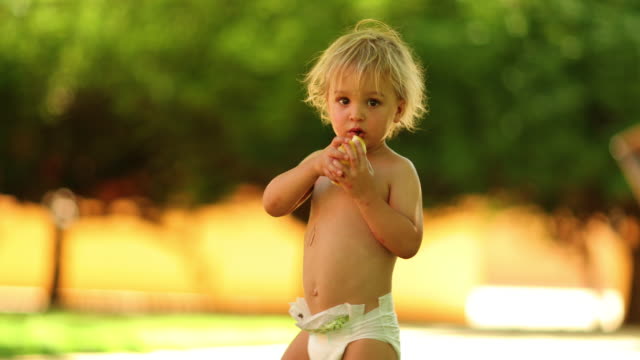 Candid-shot-of-blonde-infant-toddler-boy-with-diapers-outdoors-in-4k-clip-resolution.-Toddler-infant-child-holding-a-pear-fruit-outside