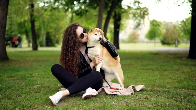 Glückliches-Mädchen-stolz-Hundebesitzer-ist-streicheln-und-küssen-ihr-Haustier-auf-dem-Rasen-im-Park-zu-sitzen,-während-das-Tier-Liebe-und-Fürsorge-genießt.-Grüne-Bäume-und-Rasenflächen-sind-sichtbar.
