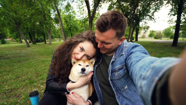 Young-wife-and-husband-are-taking-selfie-with-adorable-dog-kissing-and-hugging-each-other-and-the-animal.-Point-of-view-shot-of-happy-people,-pet-and-green-park.