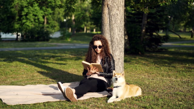 Attraktive-Studentin-liest-Buch-sitzen-auf-Plaid-unter-Baum-im-Stadtpark-mit-ihrem-Hund-liegen-in-der-Nähe-und-Sonnenlicht-zu-genießen.-Hobby,-Freizeit-und-Tiere-Konzept.