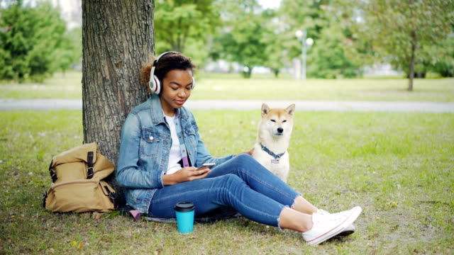 Modern-African-American-girl-is-listening-to-music-with-headphones-and-using-smartphone-relaxing-in-city-park-with-pet-dog,-stroking-and-caressing-the-animal.