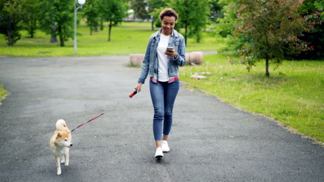 Ziemlich-afroamerikanische-Mädchen-Denim-Jacke-und-Jeans-ist-ihre-reinrassigen-Hund-spazieren-gehen,-im-Stadtpark-und-mit-Smartphone-gehen-weg-mit-Rasen-und-Bäumen-sichtbar.