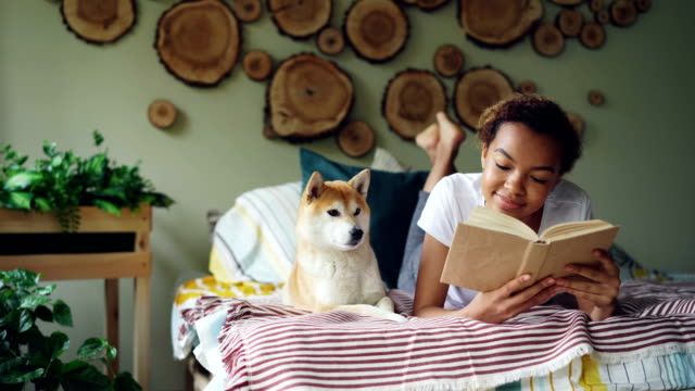 Fröhliche-Mischlinge-Teenager-Mädchen-liest-Buch-Literatur-genießen-dann-streichelte-ihr-Shiba-Inu-Hund-auf-Bett-in-ihrer-Nähe.-Hobby,-modernen-Lifestyle-und-Tiere-Konzept.