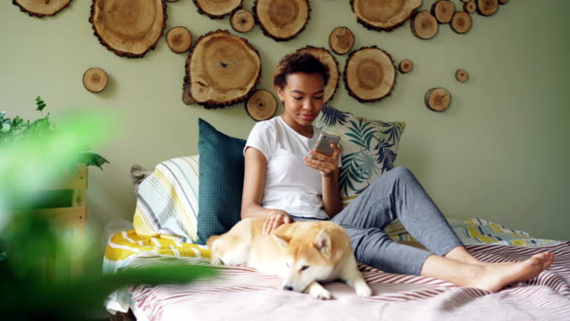 Pretty-young-woman-is-using-smartphone-and-caressing-lovely-puppy-lying-on-bed-near-its-owner-in-modern-style-bedroom-at-home.-Technology,-animals-and-people-concept.