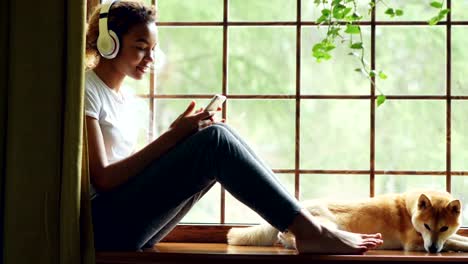 Pretty-African-American-girl-is-listening-to-music-with-headphones-and-using-smartphone-sitting-on-window-sill-with-lovely-well-bred-dog-lying-near-her.