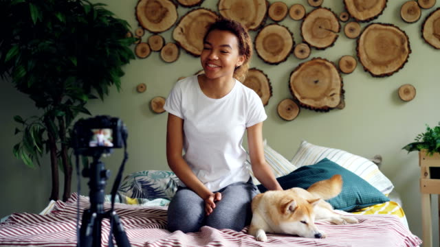 Cute-African-American-teenager-blogger-is-recording-videoblog-sitting-on-bed,-stroking-lovely-pet-dog-and-talking-to-subscribers.-Social-media-and-people-concept.