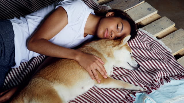 Beautiful-African-American-teenager-and-adorable-pet-dog-are-sleeping-together-on-wooden-bed,-girl-is-wearing-comfortable-pajamas-and-hugging-animal.