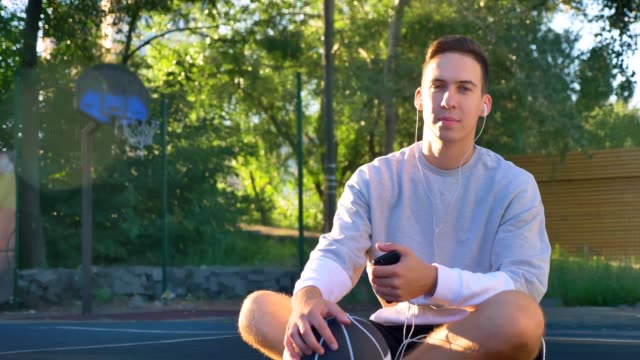 Confident-young-man-sitting-on-basketball-court-and-listening-music,-looking-at-camera,-holding-phone-and-ball,-park-in-background