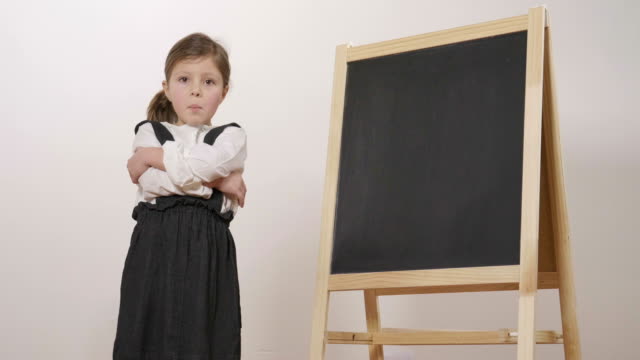 A-happy-girl-dressed-as-a-teacher-in-front-of-a-small-blackboard-holds-her-arms-folded-and-smiles.